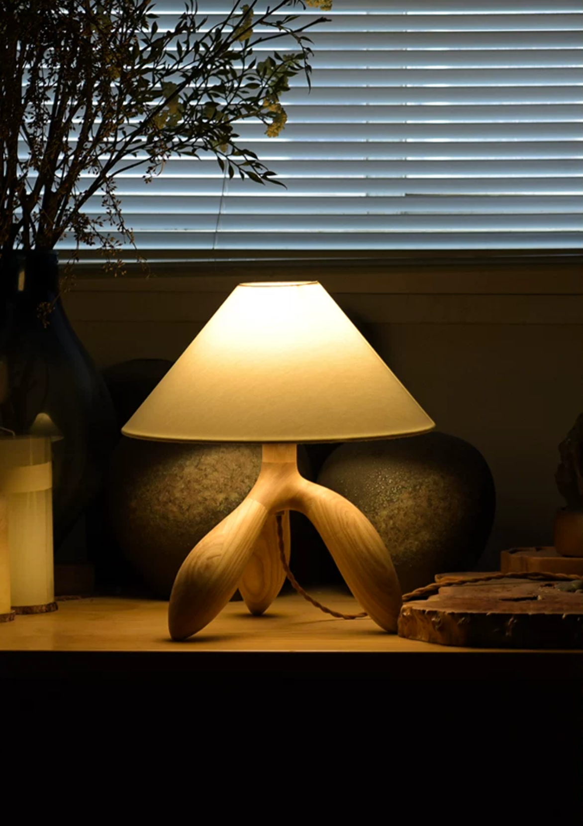 In front of venetian blinds, this dark wood Clérachic lamp glows beside potted plants on a wood table.