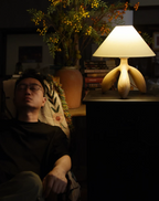 Beside a sleeping figure on a side table, this dark wood Clérachic lamp glows warm next to a leafy plant.