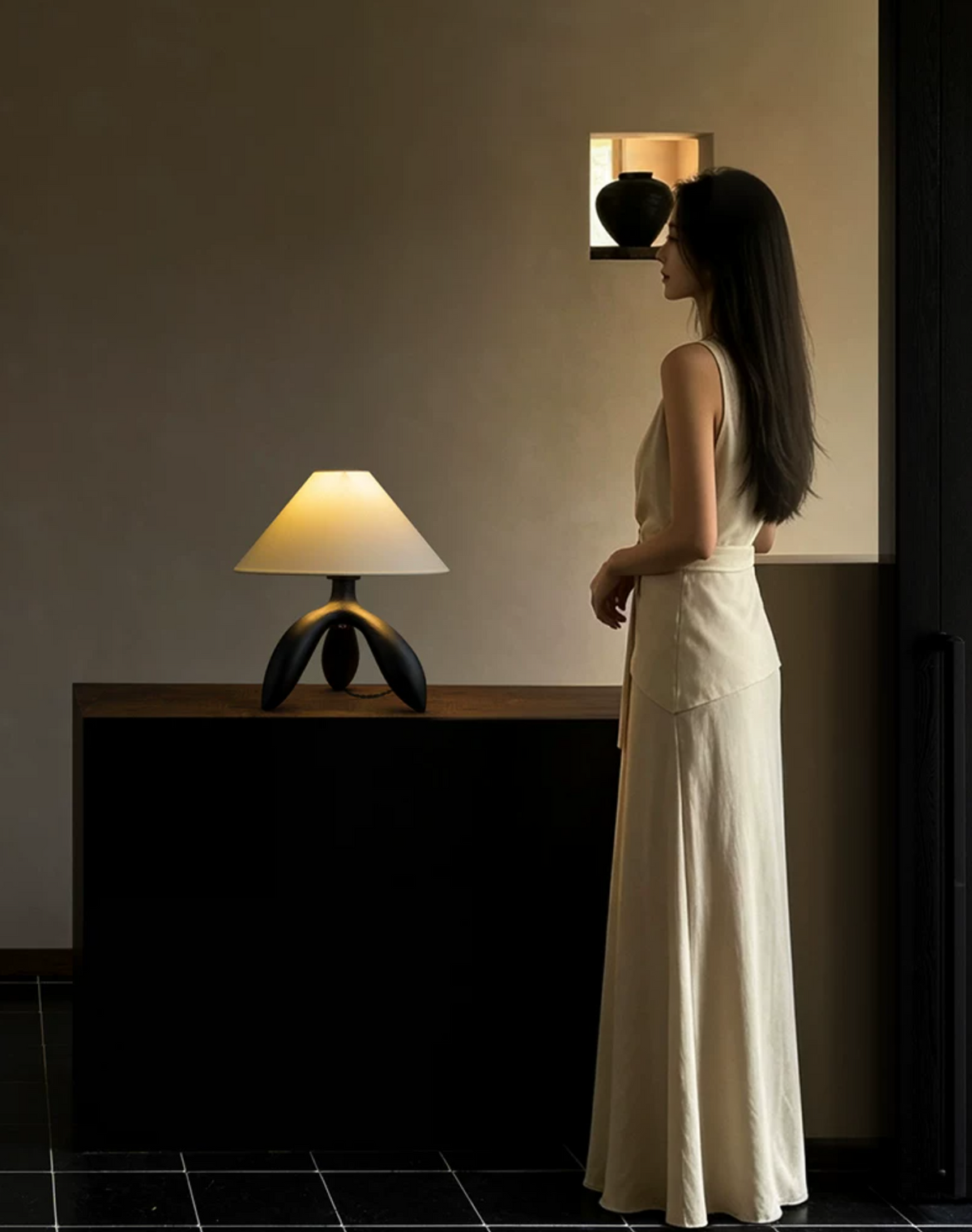 Lit on a dark cabinet, this dark wood Clérachic lamp glows beside a woman standing near a small wall shelf.