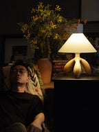 Near a sleeping figure and a leafy plant, this light wood Clérachic lamp glows atop a side table stacked with books.