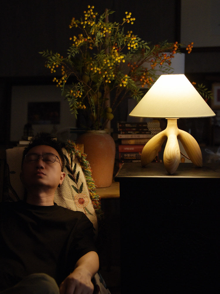 Near a sleeping figure and a leafy plant, this light wood Clérachic lamp glows atop a side table stacked with books.