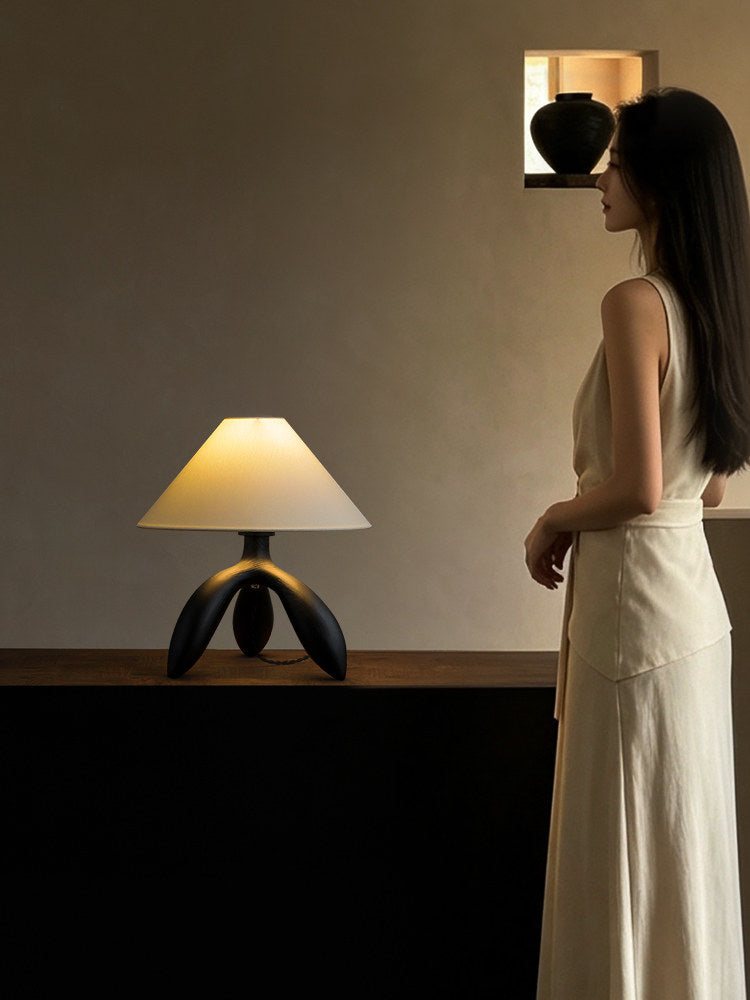 A woman in a white dress walks past this dark wood Clérachic lamp, lit on a cabinet below a small shelf.