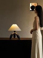 A woman in a white dress walks past this dark wood Clérachic lamp, lit on a cabinet below a small shelf.