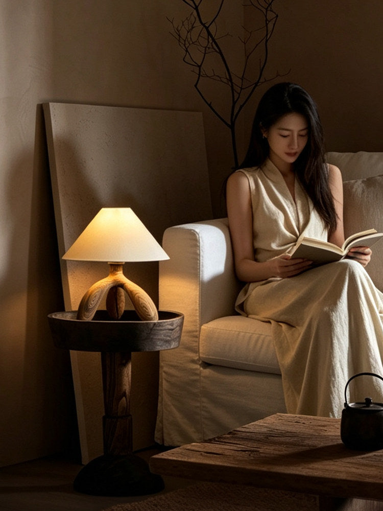 Beside an armchair and a small teapot, this dark wood Clérachic lamp glows warm while someone reads nearby.