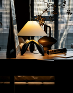 Near a shuttered window, this dark wood Clérachic lamp glows on a dark desk beside a bronze vase and rolled papers.