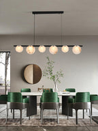 Lit above a dining table with green velvet chairs, a round brass mirror on the wall behind.