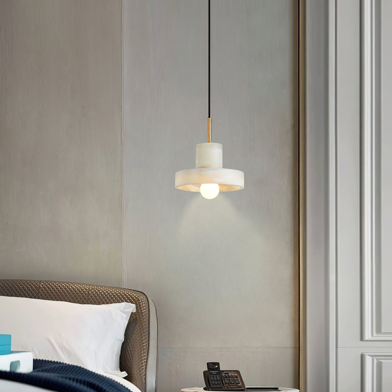Ritta pendant glowing beside a quilted headboard, its bulb throwing a soft cone onto the wall.