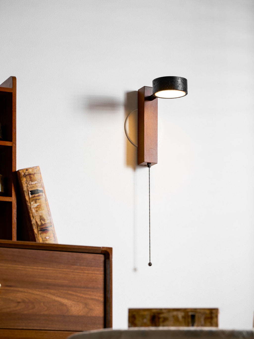 Swivelled toward the camera, this Clérachic wall light hangs its pull chain beside a wooden bookcase and round table.