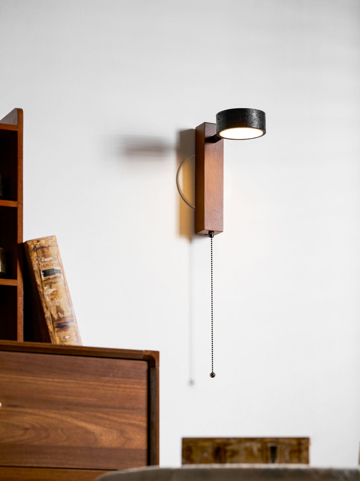 Swivelled toward the camera, this Clérachic wall light hangs its pull chain beside a wooden bookcase and round table.