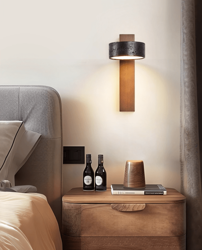 An upholstered grey headboard and a nightstand with bottles and a cup sit beneath this Clérachic wall light.