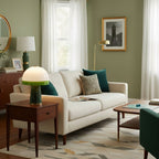 A Clérachic table lamp with a green glass rim lights a wooden side table beside a cream sofa in a sage-green living room.