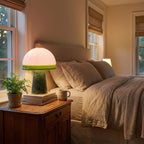 A Clérachic dome table lamp glows on a wooden nightstand beside a potted fern and stacked books in a bedroom.