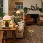 A lit Clérachic table lamp sits on a wooden side table beside a cream sofa, a fireplace and leather armchairs behind it.