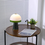 A full view of the lit Clérachic table lamp on the round table, with its plant, magazine and cord all visible.