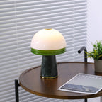 A Clérachic dome lamp sits lit on a round wooden table beside a potted plant and a magazine, blinds behind it.