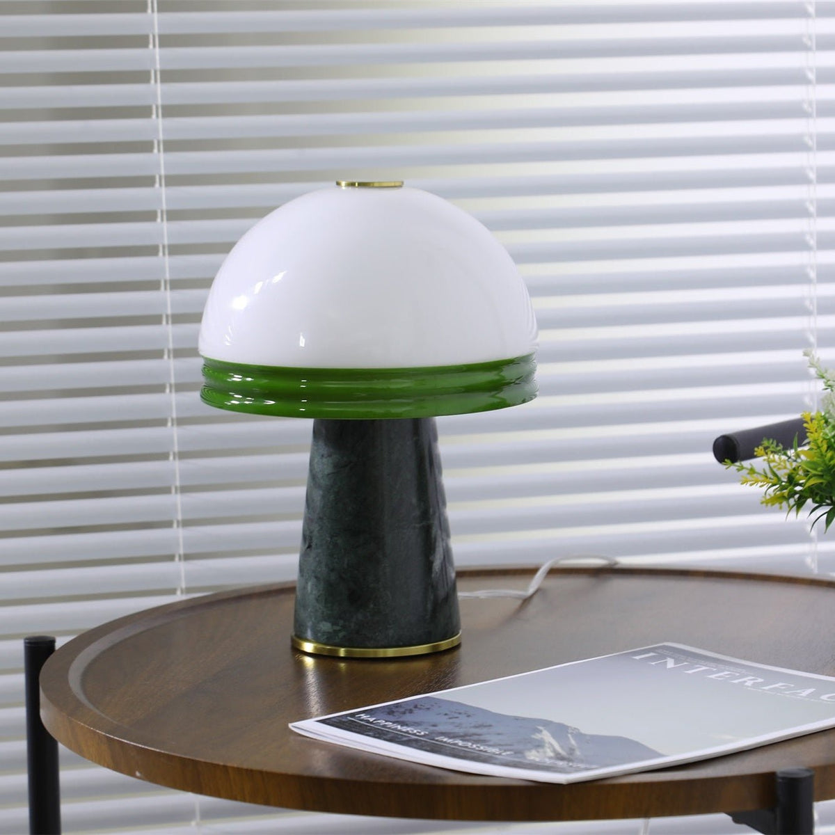 Wide view of a Clérachic table lamp on a round wooden table, its cord trailing beside an open magazine.