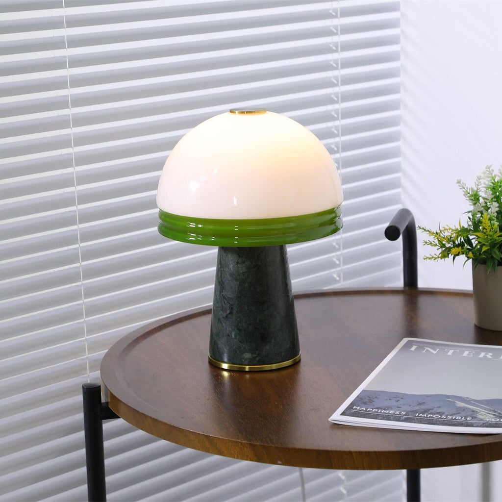 Angled view of the lit Clérachic table lamp on the round table, venetian blinds softly lit behind it.