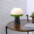 Angled view of the lit Clérachic table lamp on the round table, venetian blinds softly lit behind it.
