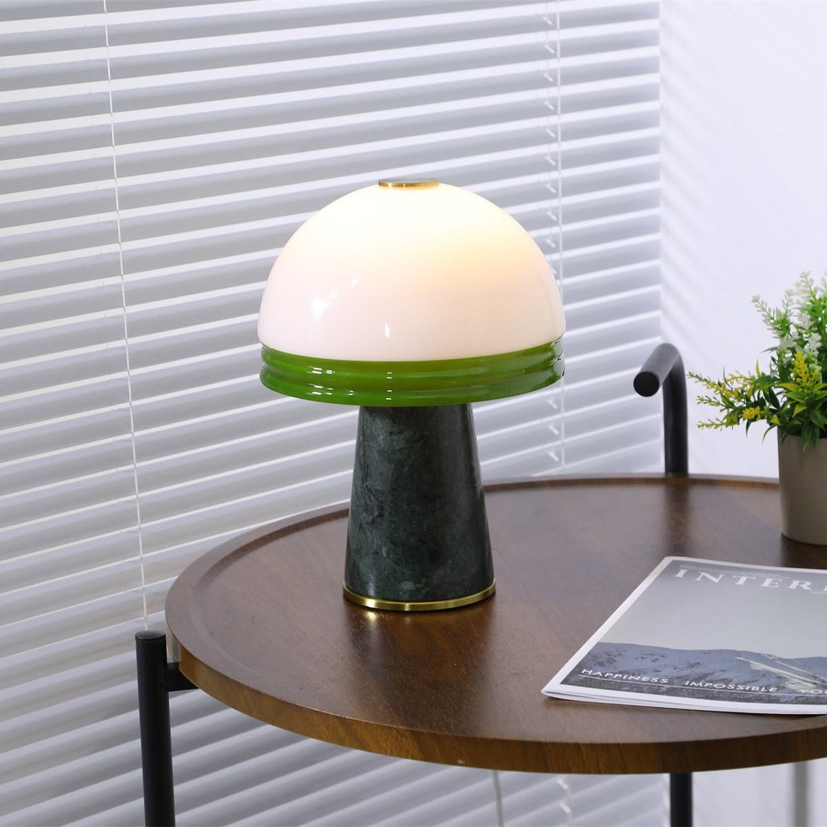 Angled view of the lit Clérachic table lamp on the round table, venetian blinds softly lit behind it.