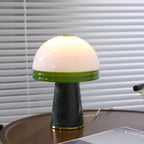 Close-up of a Clérachic table lamp's white glass dome and green rim on a round wooden table against venetian blinds.