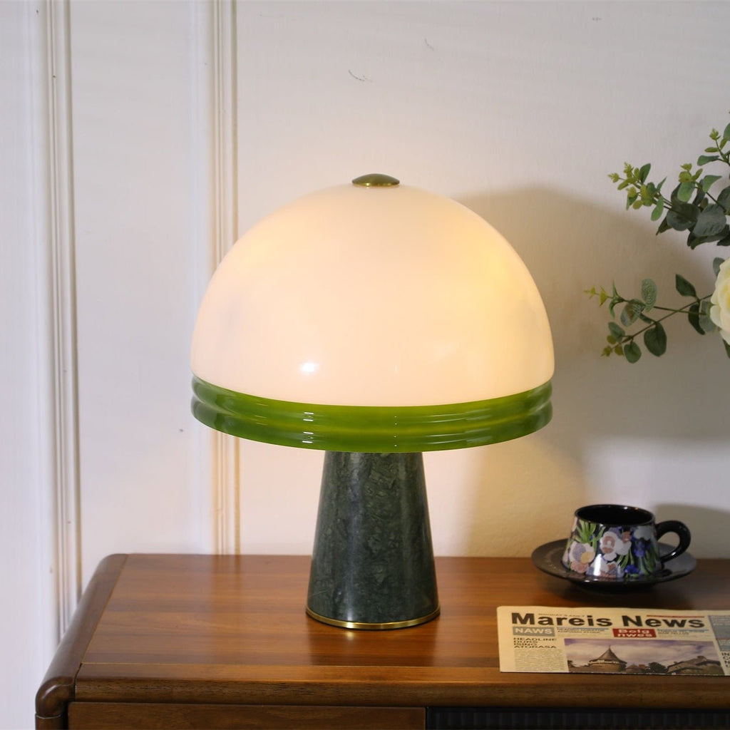 Centred in frame on a wood cabinet, the Clérachic dome table lamp glows beside a teacup, saucer and a folded newspaper.