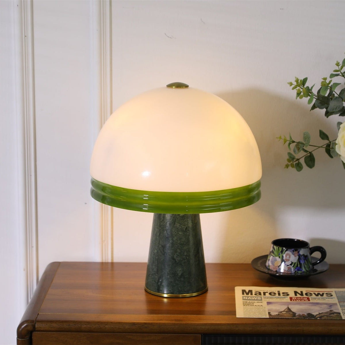 Centred in frame on a wood cabinet, the Clérachic dome table lamp glows beside a teacup, saucer and a folded newspaper.
