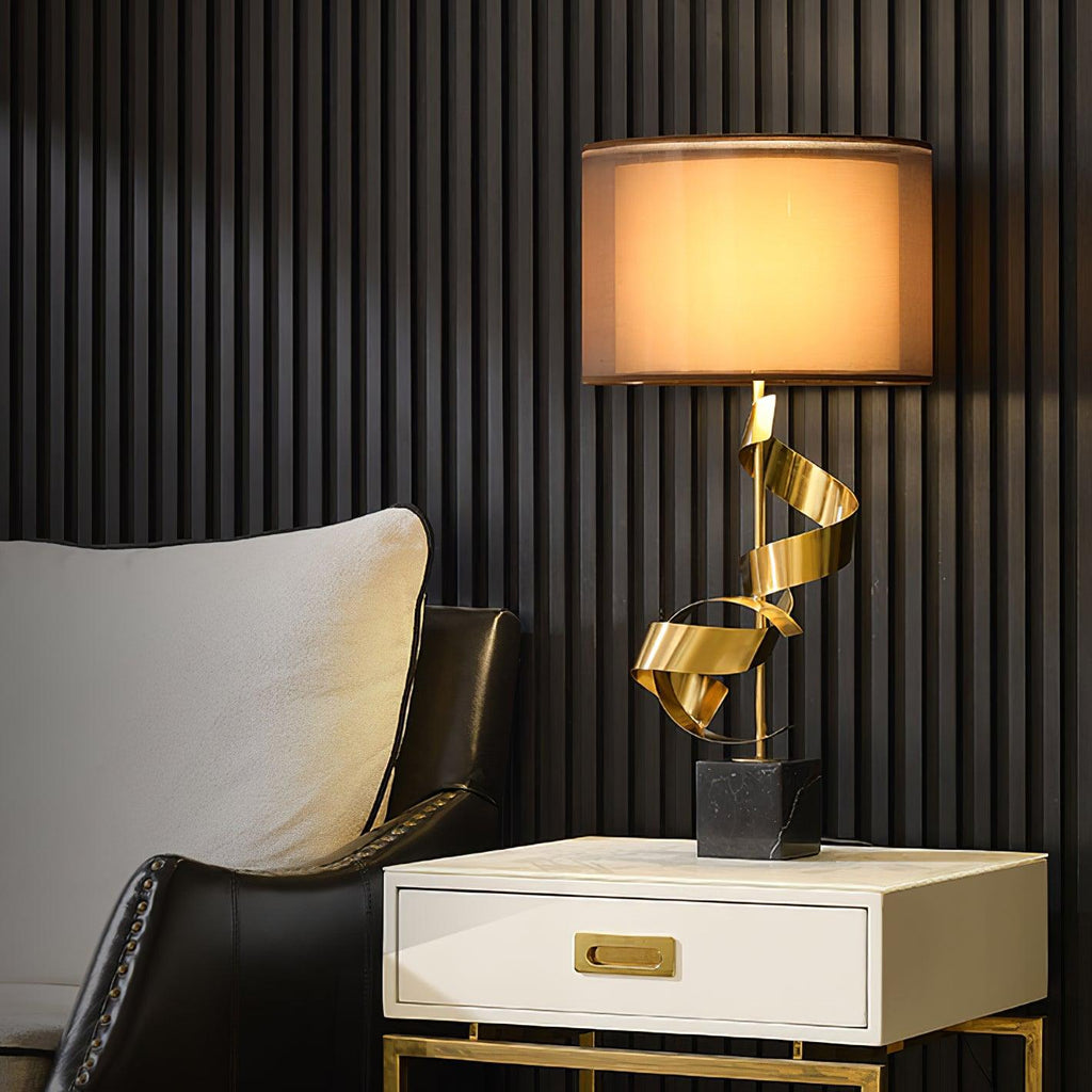 Clérachic gold spiral table lamp glowing on a white bedside drawer unit, a dark ribbed wall and leather chair behind.