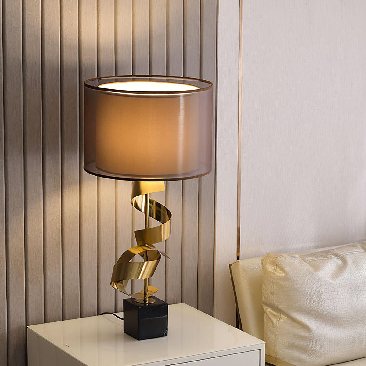 Clérachic gold spiral table lamp lit on a white bedside nightstand, its warm glow softening the fluted wall panelling.