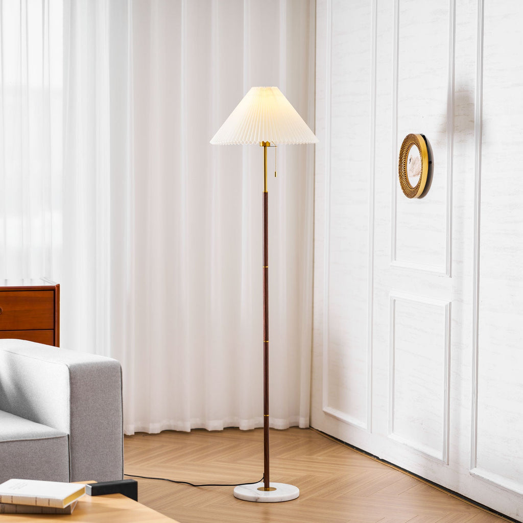 A Clérachic pleated-shade floor lamp lit in a living room corner, beside a grey armchair, sheer curtains and a round wall mirror.