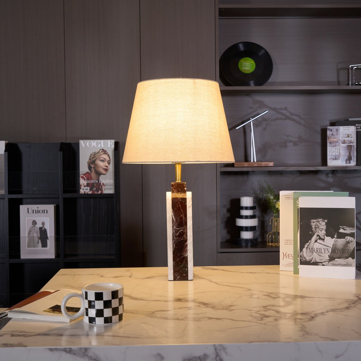The Clérachic marble table lamp on a stone side table, its beige shade lit warmly in front of a dark bookshelf.