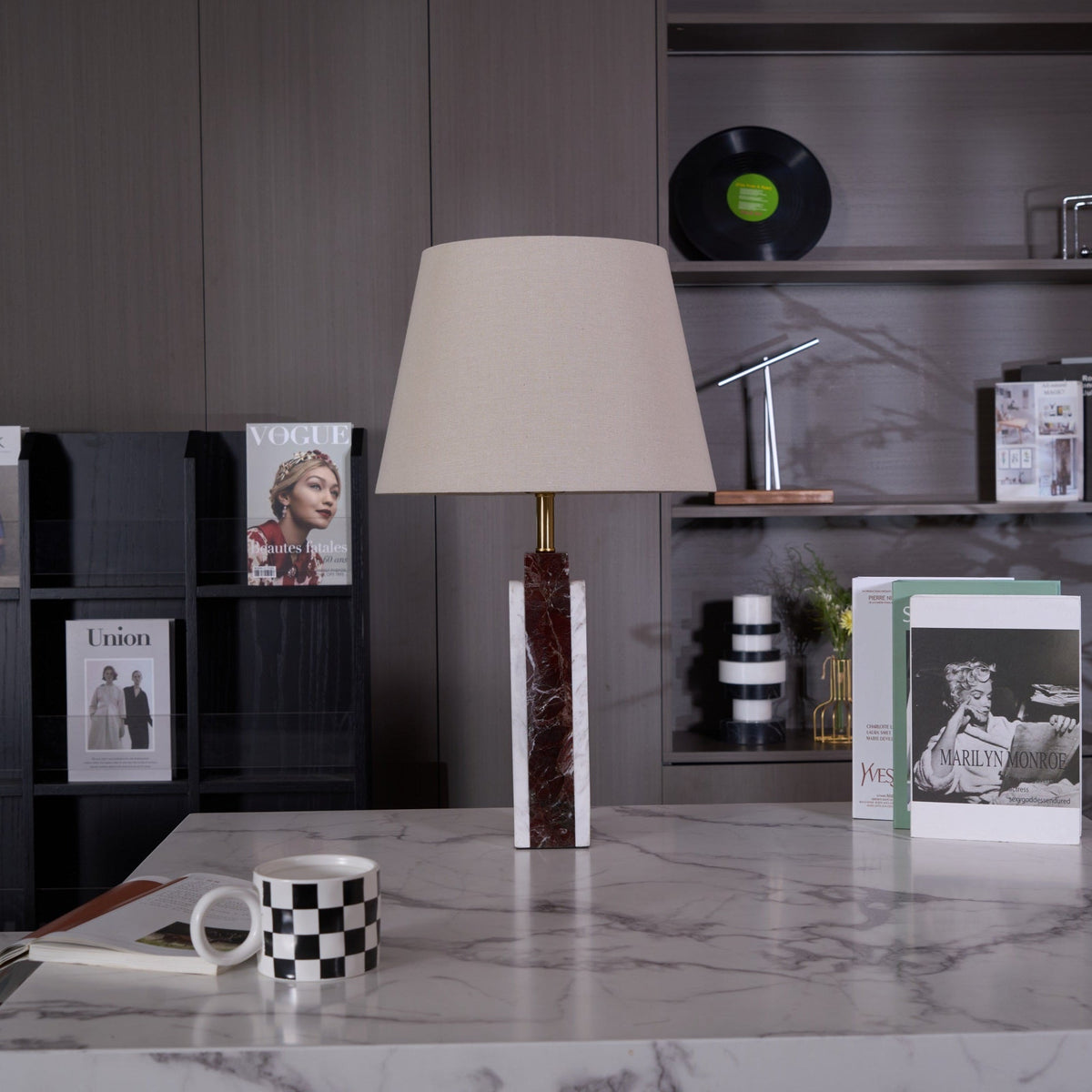 A Clérachic table lamp lit on a stone plinth, a magazine and checkerboard mug arranged beside its dark veined base.