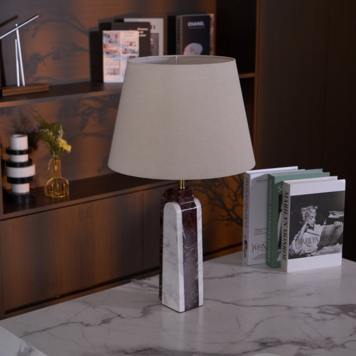 The Clérachic marble table lamp sits unlit on a stone plinth, its shade reading pale grey against dark wood shelving.