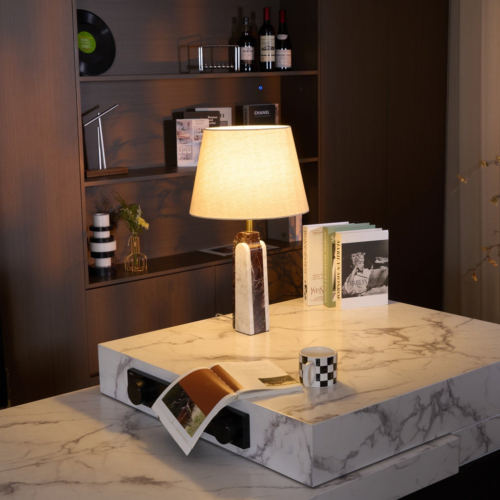 A Clérachic marble table lamp lit on a stone side table, framed by a dark bookshelf holding records and wine bottles.