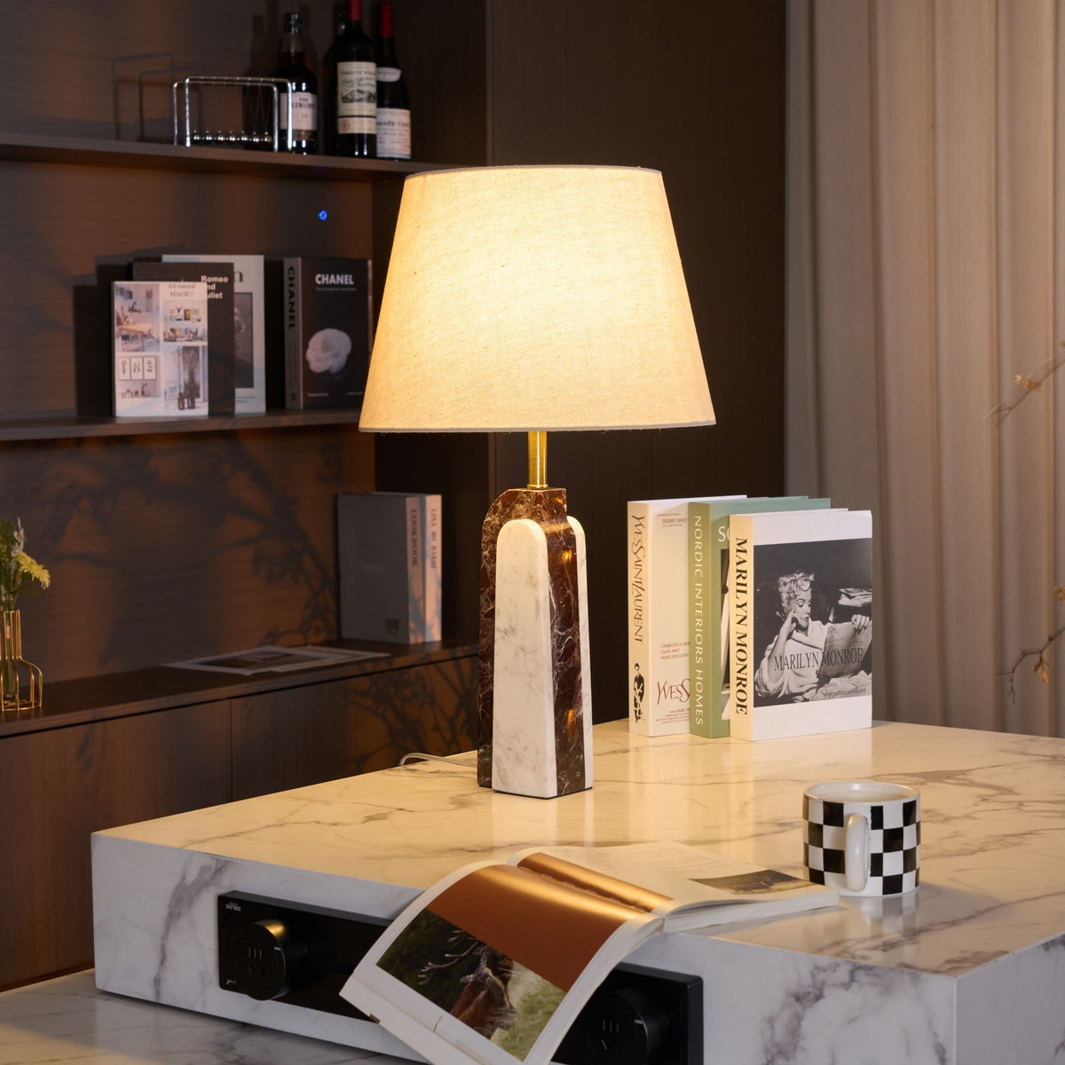 The Clérachic marble table lamp lit on a white stone plinth, a magazine draped over the edge beside a checkerboard mug.