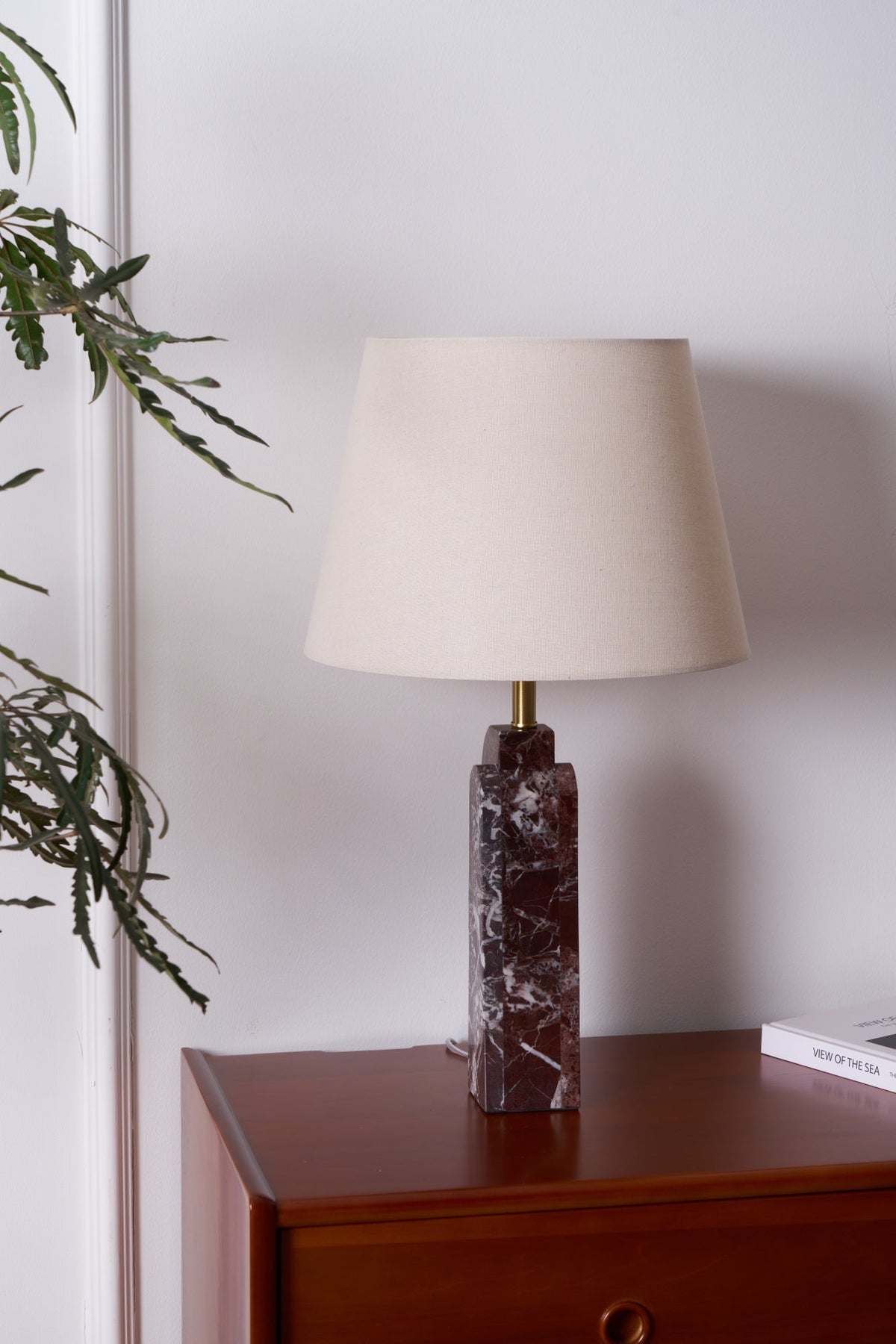 A cropped view of the Clérachic table lamp lit on a dresser, a potted plant visible at the edge of the frame.
