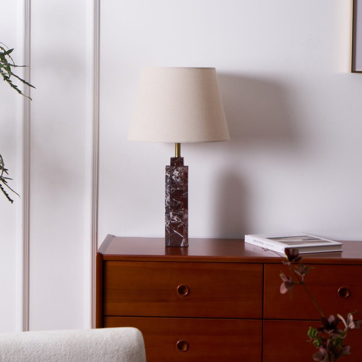 A Clérachic table lamp stands on a dresser against a plain wall, shade unlit, one book resting at its base.
