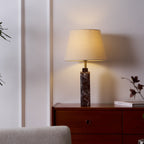 A Clérachic marble table lamp lit beside a cream armchair, its dark veined base on a walnut dresser with dried flowers.
