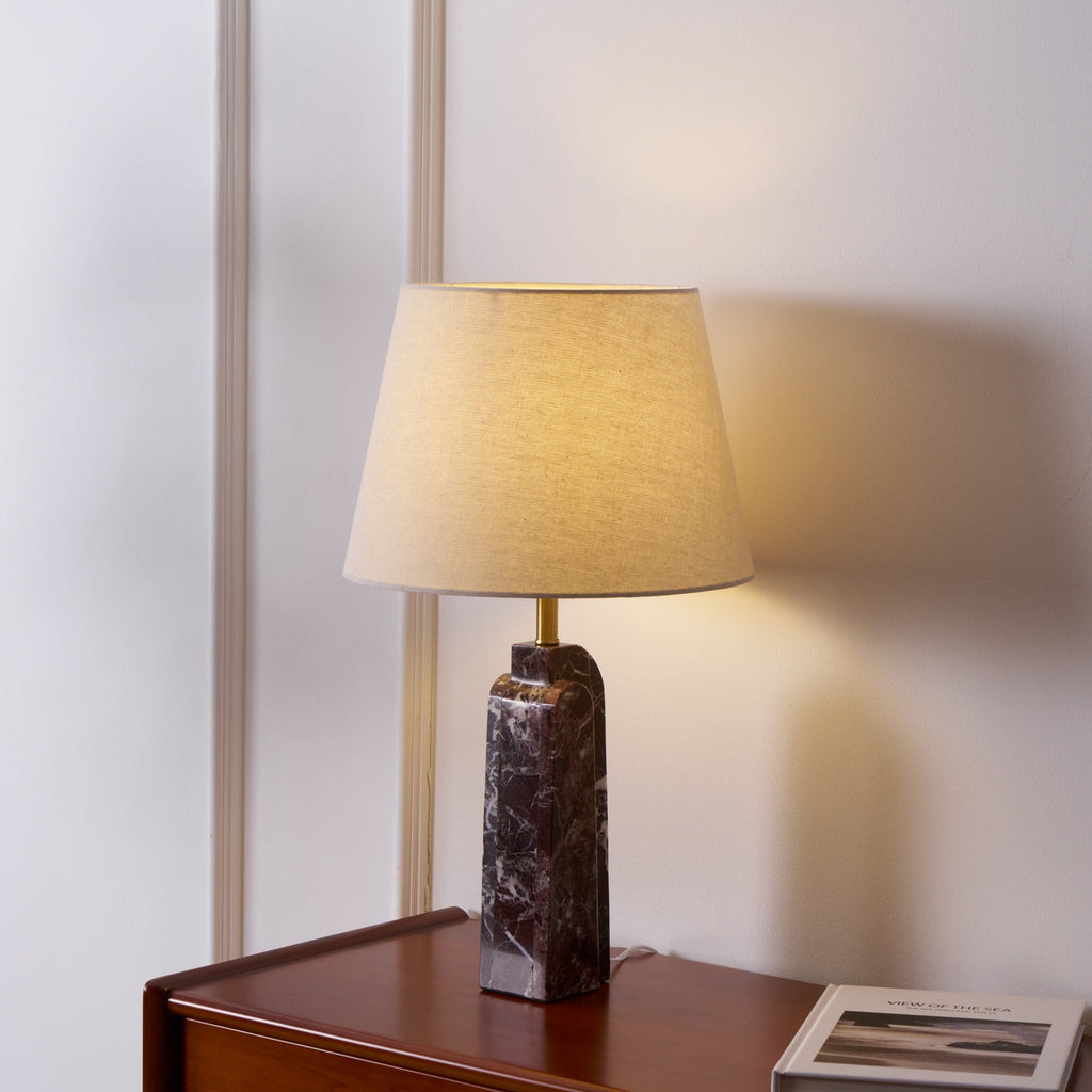 The Clérachic marble table lamp lit on a bedside dresser, shade angled to show the linen weave against the warm glow.