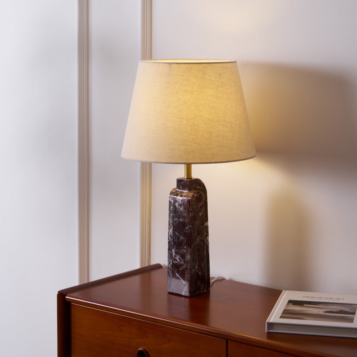 A tall, narrow view of the Clérachic marble table lamp lit on a bedside dresser, its beige shade filling the frame.