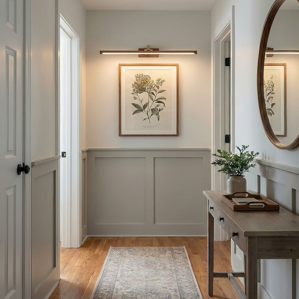 A botanical print hangs beneath this Clérachic wall light in brass, near a round mirror in a pale hallway.