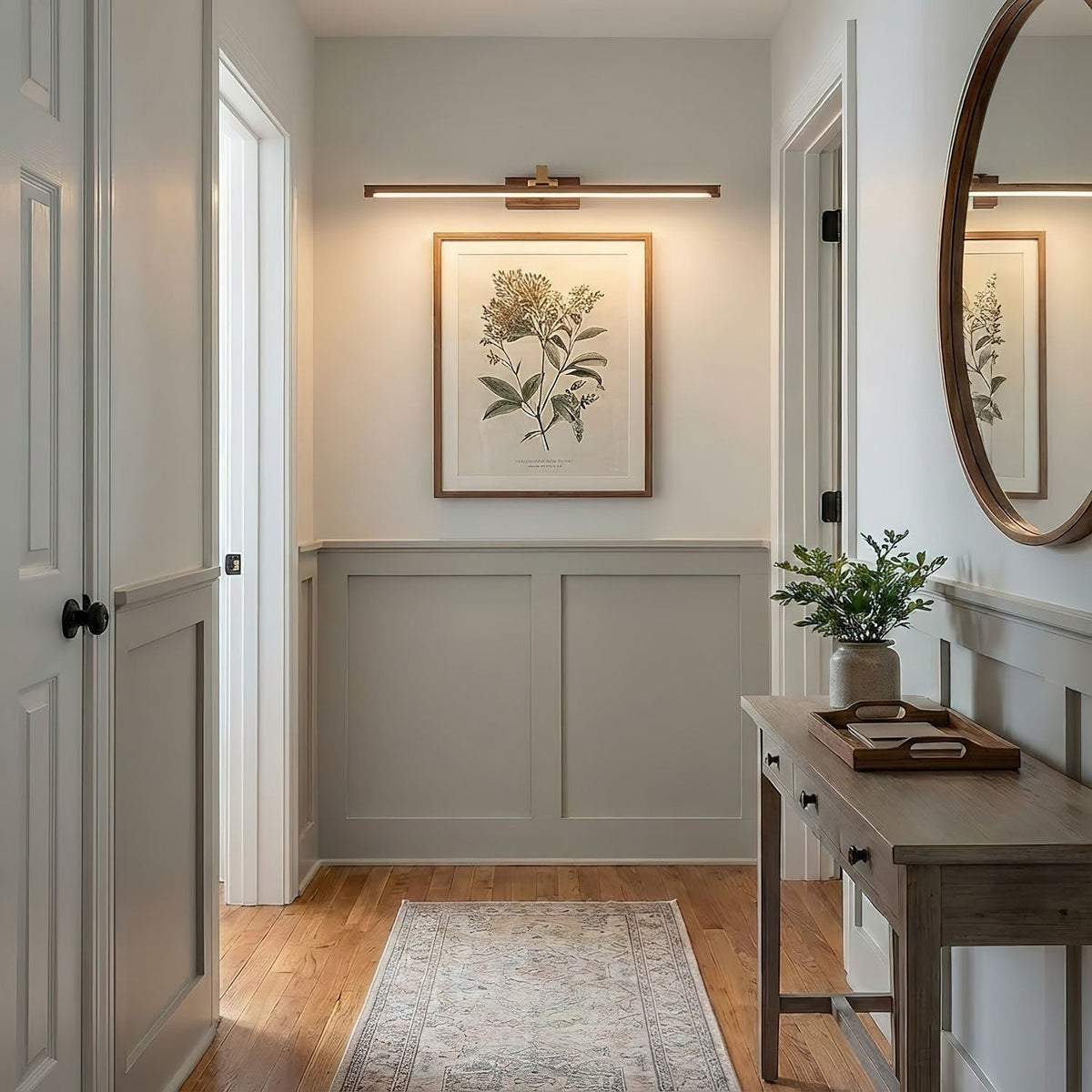 A botanical print hangs beneath this Clérachic wall light in brass, near a round mirror in a pale hallway.