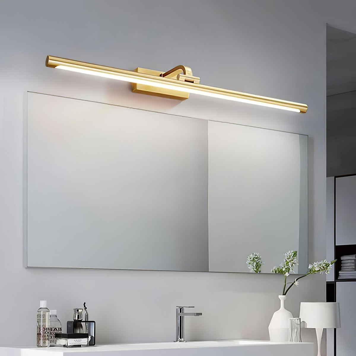 A bathroom vanity with fresh flowers sits beneath this Clérachic wall light in brass, lit, above a wide mirror.