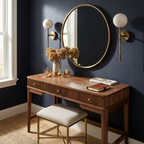 Two brass Clérachic wall sconces flank an oval mirror above a wood vanity desk with dried flowers.