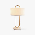 A Clérachic table lamp with a curved brass loop frame and white linen drum shade stands on a pale marble base against a plain light backdrop.