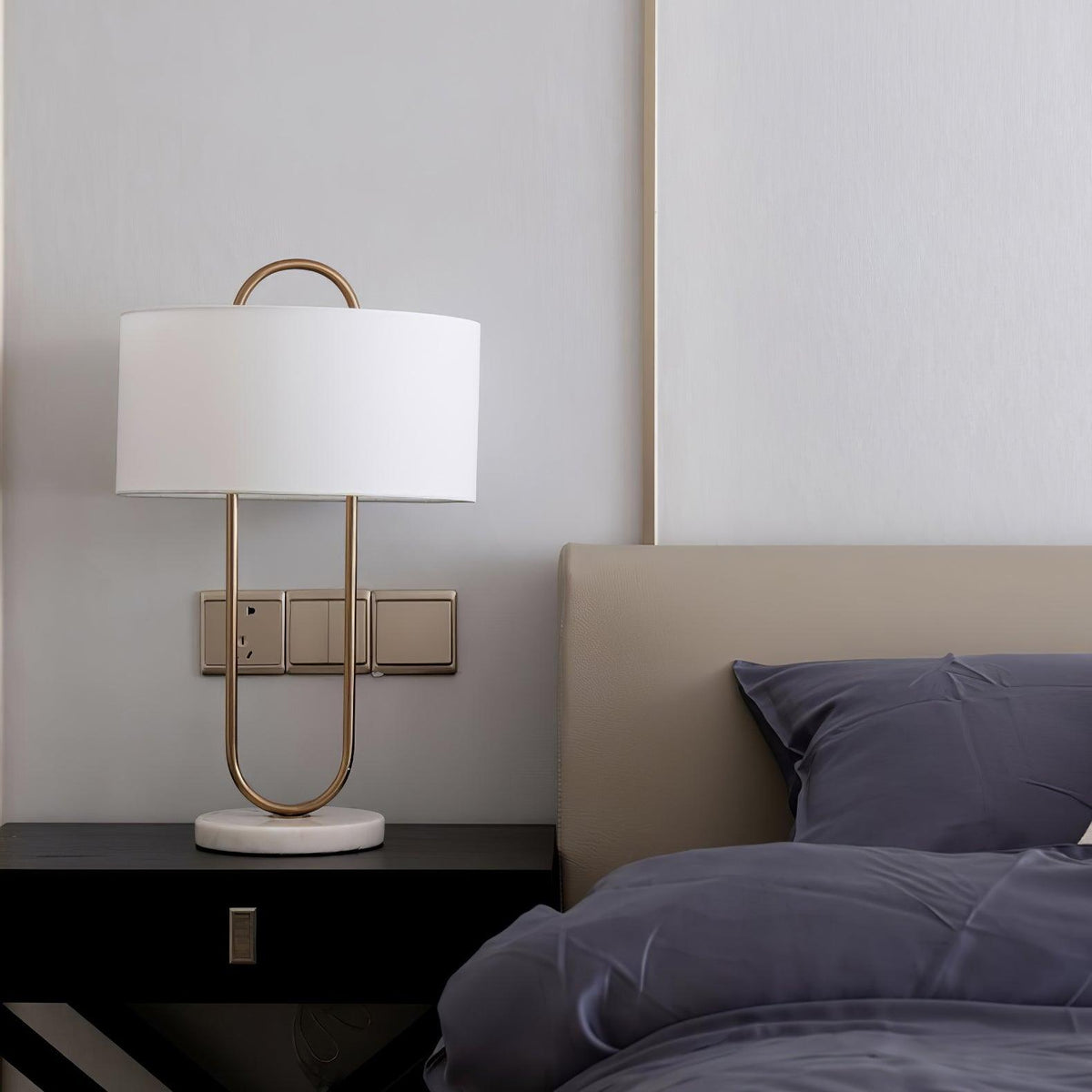 Beside a taupe headboard and navy bedding, this Clérachic table lamp's gold frame stands close to a wall switch plate on the nightstand.