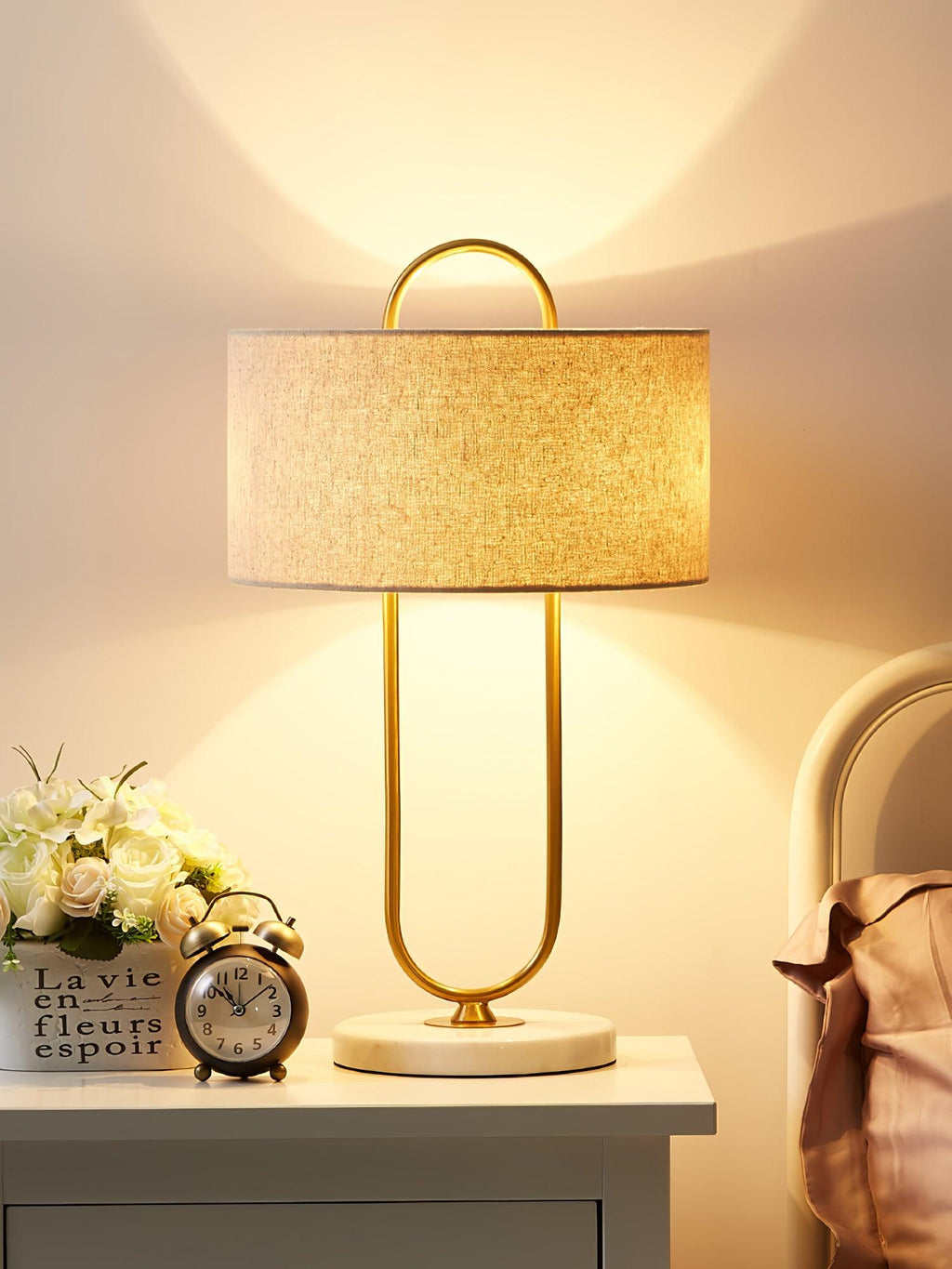 Lit warm gold, this Clérachic table lamp glows beside a vintage alarm clock and a small vase of flowers on a cream bedside table.