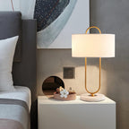 On a grey bedside table beside a tufted headboard, this Clérachic lamp's linen shade and gold frame sit near a small tray and framed art.