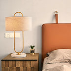 Beside a tan leather headboard, this Clérachic lamp's linen shade catches warm light next to a small potted plant and a wall switch plate.