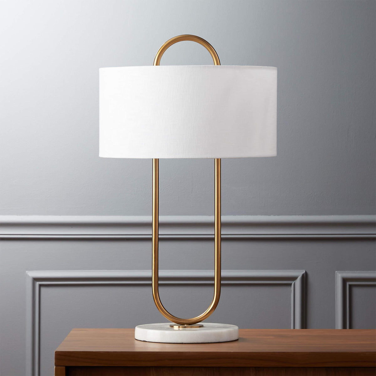 Centred on a wood table against a light grey panelled wall, this unlit Clérachic table lamp shows its full brass frame and marble base.