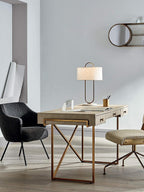 On a gold-framed desk between two dark chairs, this Clérachic table lamp sits lit in a bright, minimal home office setting.